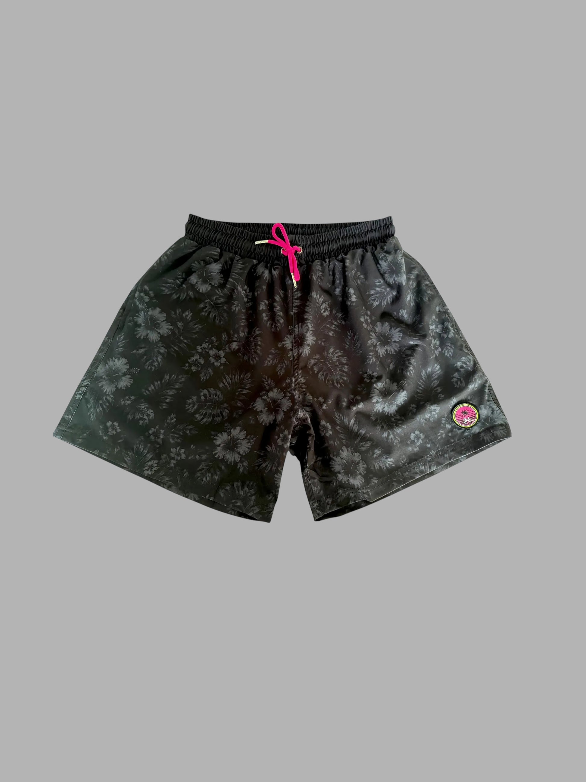 Black athletic shorts with a subtle tonal floral pattern, featuring a bright pink drawstring and a small pink circular logo patch on the left leg, displayed against a solid gray background.